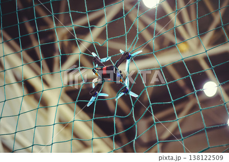 A horizontal shot of a small FPV racing drone with blue propellers stuck in a green safety net during an indoor flight. A horizontal shot of a small FPV racing drone with blue propellers stuck in a green safety net during an indoor flight. 138122509