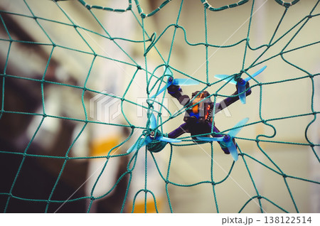 A horizontal shot of a small FPV racing drone with blue propellers stuck in a green safety net during an indoor flight. 138122514