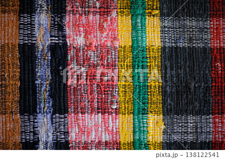 A horizontal top-down shot of a traditional handmade woven rug. The rug features vertical stripes in various colors, including red, yellow, green, black, and blue, with visible fabric textures. A horizontal top-down shot of a traditional handmade woven rug. The rug features vertical stripes in various colors, including red, yellow, green, black, and blue, with visible fabric textures. 138122541