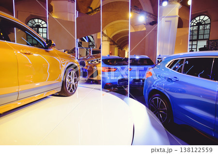 A wide horizontal shot of a car exhibition featuring golden and blue luxury vehicles. The composition is framed by vertical neon light pillars, creating a futuristic atmosphere in an arched hall. 138122559