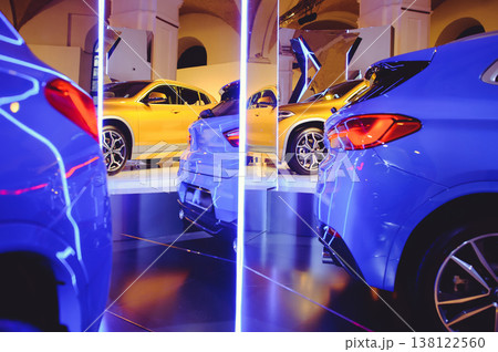 A wide-angle horizontal shot of a luxury car dealership, featuring a row of premium golden and blue vehicles parked under a series of illuminated arches and neon vertical pillars. 138122560