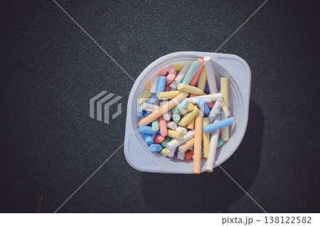 A horizontal top-down shot of a white plastic bowl filled with numerous pieces of multi-colored sidewalk chalk, placed on a dark asphalt surface. 138122582