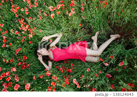 A horizontal high-angle shot of a young woman with long dark hair, wearing a red patterned summer dress, lying gracefully among green grass and vibrant blooming red poppies A horizontal high-angle shot of a young woman with long dark hair, wearing a red patterned summer dress, lying gracefully among green grass and vibrant blooming red poppies 138124043