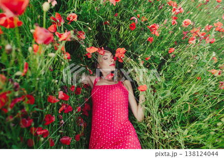 A horizontal high-angle shot of a young woman with long dark hair, wearing a red patterned dress, lying peacefully with closed eyes among tall green grass and blooming red poppies. A horizontal high-angle shot of a young woman with long dark hair, wearing a red patterned dress, lying peacefully with closed eyes among tall green grass and blooming red poppies. 138124044