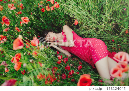 A horizontal shot of a young woman with long dark hair, wearing a red patterned dress, lying on her side with closed eyes as if sleeping among lush green grass and blooming red poppies. A horizontal shot of a young woman with long dark hair, wearing a red patterned dress, lying on her side with closed eyes as if sleeping among lush green grass and blooming red poppies. 138124045