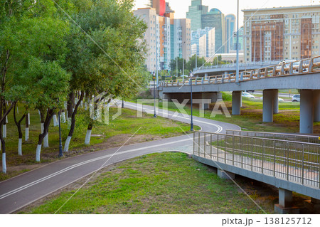 Bicycle path in city park winding under highway overpass. Pedestrian walkway near concrete flyover with metal railing. Urban landscape with road infrastructure and green trees in city. 138125712
