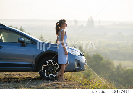 Happy young woman in blue dress standing near her vehicle looking at sunset view of summer nature. Travelling and vacation concept. Happy young woman in blue dress standing near her vehicle looking at sunset view of summer nature. Travelling and vacation concept. 138127496