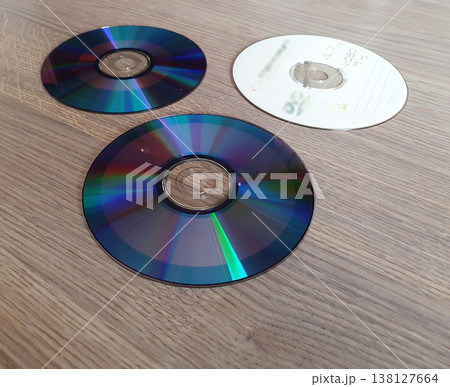 Three compact discs on a wooden surface. One white disc and two with rainbow reflection. Retro media of information, music and video games. Three compact discs on a wooden surface. One white disc and two with rainbow reflection. Retro media of information, music and video games. 138127664