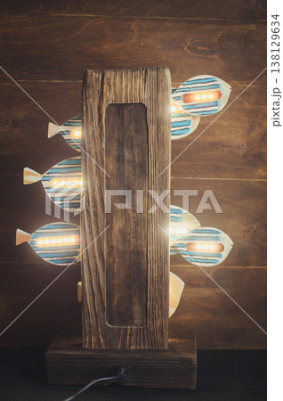 Rear view of a handcrafted wooden lamp with LED-lit striped fish elements on a rustic timber base in a dark setting. 138129634