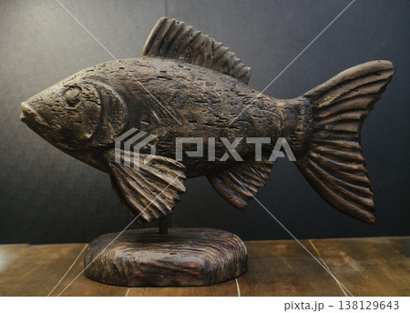 Hand-carved wooden fish sculpture with aged, weathered texture on a solid timber stand against a dark background. 138129643