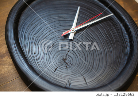 Close-up perspective of a handcrafted wall clock made from a scorched black wood slice, emphasizing the rhythmic circular annual rings and silver hands. 138129662