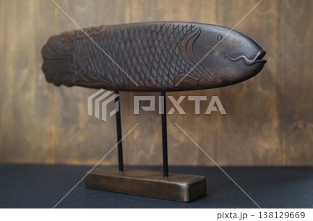 Perspective view of a hand-carved wooden fish sculpture on a minimalist black metal stand, showcasing the detailed head, geometric scales, and flowing tail patterns. Perspective view of a hand-carved wooden fish sculpture on a minimalist black metal stand, showcasing the detailed head, geometric scales, and flowing tail patterns. 138129669