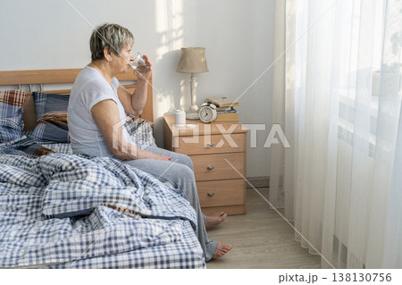 Senior woman taking a drink of water in her bedroom, sitting on the bed. Practicing her daily health and wellness routine, medication routine, prevention of age-related diseases Senior woman taking a drink of water in her bedroom, sitting on the bed. Practicing her daily health and wellness routine, medication routine, prevention of age-related diseases 138130756