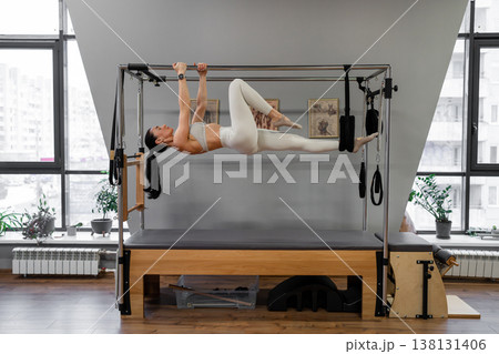 Young woman laying on pilates reformer with legs up, performing pilates exercises indoors. Pilates exercise, laying, reformer, fitness, indoor. 138131406