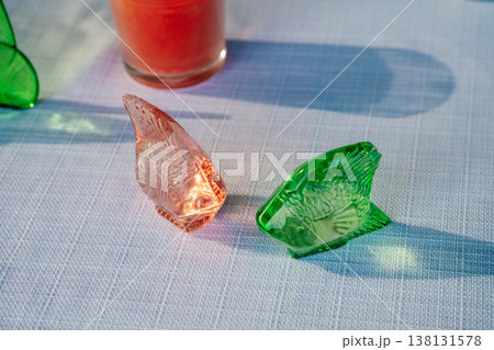 Colored Glass Leaves On Sunlit Table Next To Tall Red Drink, Sharp Shadows, Textured Linen Surface Colored Glass Leaves On Sunlit Table Next To Tall Red Drink, Sharp Shadows, Textured Linen Surface 138131578