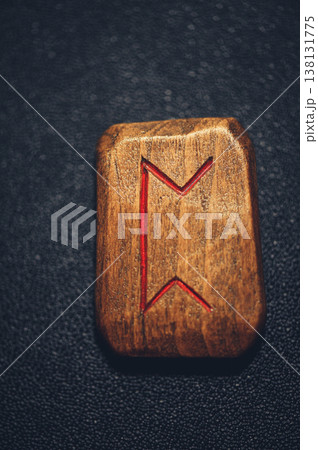 A vertical close-up of a handcrafted wooden Perthro rune stone, featuring the carved symbol filled with red pigment on a rustic grain. 138131775