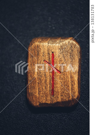 Vertical close-up of a handcrafted wooden Nauthiz rune stone, featuring the carved symbol filled with red pigment on a rustic grain. Vertical close-up of a handcrafted wooden Nauthiz rune stone, featuring the carved symbol filled with red pigment on a rustic grain. 138131783