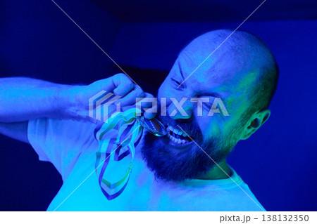 Male Winner Biting Medal in Blue Light Celebrating Victory and Triumph With Joyful Expression 138132350