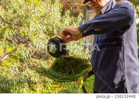 A man cleans grass trimmer from grass. Gardener maintaining backyard with string trimmer. Seasonal landscaping tool for farm maintenance and land cleanup work. 138132448