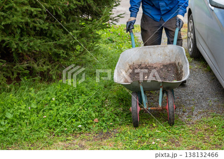 Gardener man pushing empty wheelbarrow or metal cart in garden. Agricultural equipment for horticulture, landscaping, landscaping maintenance and seasonal work. Gardener man pushing empty wheelbarrow or metal cart in garden. Agricultural equipment for horticulture, landscaping, landscaping maintenance and seasonal work. 138132466
