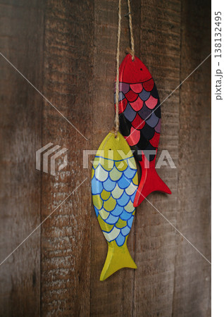 Vertical close-up of two handcrafted wooden fish sculptures, one yellow and one red with painted scales, hanging by twine against a rustic wooden wall. Vertical close-up of two handcrafted wooden fish sculptures, one yellow and one red with painted scales, hanging by twine against a rustic wooden wall. 138132495