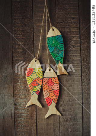 Vertical view of three handcrafted wooden fish sculptures with vibrant painted patterns on a natural wood base, hanging on twine against a rustic plank wall. 138132498