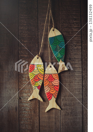 Vertical view of three handcrafted wooden fish sculptures with vibrant painted patterns on a natural wood base, hanging on twine against a rustic plank wall. 138132499