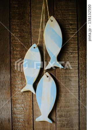 A vertical view of three handcrafted wooden fish sculptures with a distressed light blue and white stripe pattern, hanging on twine against a rustic wooden plank wall. A vertical view of three handcrafted wooden fish sculptures with a distressed light blue and white stripe pattern, hanging on twine against a rustic wooden plank wall. 138132500