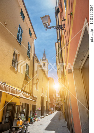 Cobblestone narrow street winding through an historic italian old town, framed by colorful buildings with shutters, a church steeple rising against the blue sky, and a bright sun flare Cobblestone narrow street winding through an historic italian old town, framed by colorful buildings with shutters, a church steeple rising against the blue sky, and a bright sun flare 138133084