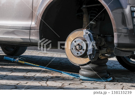 A car brake disc with visible rust and dust, mounted on a vehicle lifted by a jack. Brake caliper 138133239