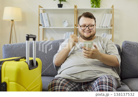 Happy man choosing travel on smartphone at home with yellow suitcase nearby 138137532