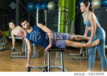 Female trainer helping young man performing side plank on pilates chair Female trainer helping young man performing side plank on pilates chair 138139764