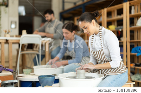 Girl craftspersons busy molding clay products on pottery wheel Girl craftspersons busy molding clay products on pottery wheel 138139890