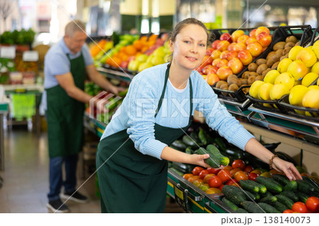 Middle-aged saleswoman putting cucumbers on food stall in greengrocery 138140073
