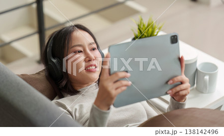 Woman using tablet and headphones while relaxing on couch 138142496
