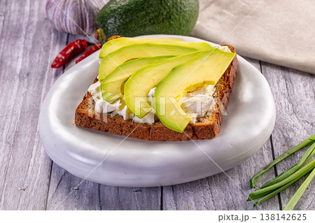 Avocado toast with soft cheese on dark rye bread. A healthy breakfast sandwich on a white plate over a rustic wooden table. 138142625