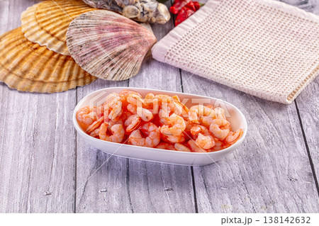 Cooked peeled shrimp in a ceramic bowl on a rustic gray wooden table with a seashell. 138142632