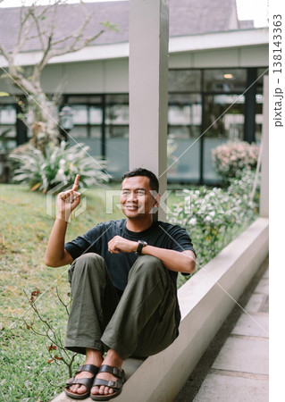 Asian Man Sitting on Low Wall Relaxing by the Garden Asian Man Sitting on Low Wall Relaxing by the Garden 138143363