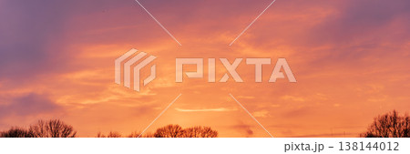 Panoramic sunset sky with fiery orange and pink clouds above tree silhouettes 138144012