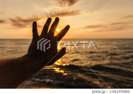 Hand reaching toward sunset over sea horizon creating warm travel lifestyle background Hand reaching toward sunset over sea horizon creating warm travel lifestyle background 138145060
