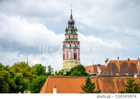 The castle tower stands tall in Cesky Krumlov surrounded by trees. The town features traditional buildings with tiled roofs under a cloudy sky. Visitors explore the streets below in summer. The castle tower stands tall in Cesky Krumlov surrounded by trees. The town features traditional buildings with tiled roofs under a cloudy sky. Visitors explore the streets below in summer. 138145974
