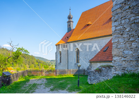 Visitors walk near Holy Trinity Church in Czechia during a sunny day. The church stands with a stone wall and red roof. Green hills surround the area, creating a scenic view for all. 138146022