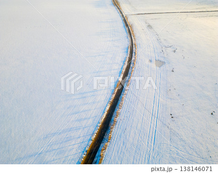 An aerial view of an asphalt road winding through a snowy field. The scene captures the contrast between the dark road and bright white snow. The photo shows winter conditions. 138146071