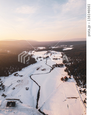 Snow covers the village of Jizerka, surrounded by trees and hills. The sun sets, casting light on the winding river and open fields, creating a winter scene in the mountains. 138146081