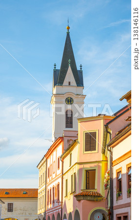 The Church of Our Lady the Queen and St. Jilj rises above colorful buildings in Trebon. The sky is clear with sunlight shining on the tower, marking an evening scene in the town. The Church of Our Lady the Queen and St. Jilj rises above colorful buildings in Trebon. The sky is clear with sunlight shining on the tower, marking an evening scene in the town. 138146116
