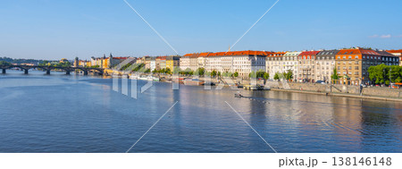 People enjoy their time near Rasn Embankment in Prague. The sun shines bright while boats move slowly on the Vltava River. Buildings line the embankment, filled with green trees. 138146148