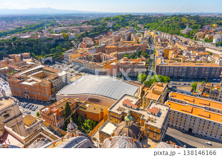 People visit Paul VI Hall in Vatican City. The area shows buildings, roads, and trees in the distance. Visitors enjoy the view and take photos of the surroundings on a sunny day. 138146166