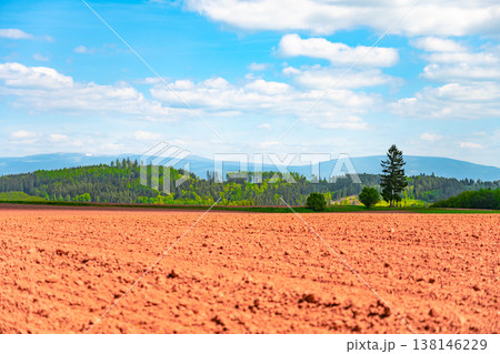 The scene shows a large area of agricultural land featuring red clay soil in the vicinity of Nova Paka, Czechia. Trees and hills are visible in the background under a bright sky. The scene shows a large area of agricultural land featuring red clay soil in the vicinity of Nova Paka, Czechia. Trees and hills are visible in the background under a bright sky. 138146229
