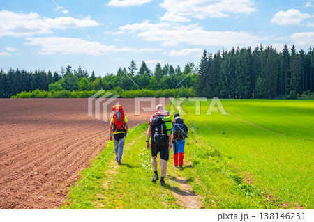 Four people walk on a path through agricultural land in Nova Paka, Czechia. Red clay soil and green fields are visible under a bright blue sky with clouds. 138146231