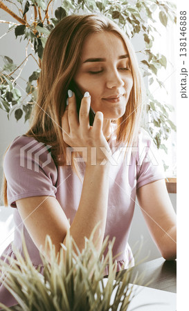 Woman talking on the phone while sitting by a window with plants around her Woman talking on the phone while sitting by a window with plants around her 138146888